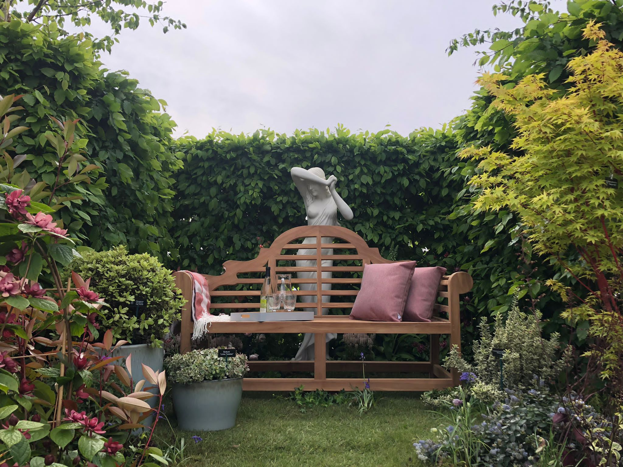 A bench with a female statue behind it in a hedge lined designer garden.